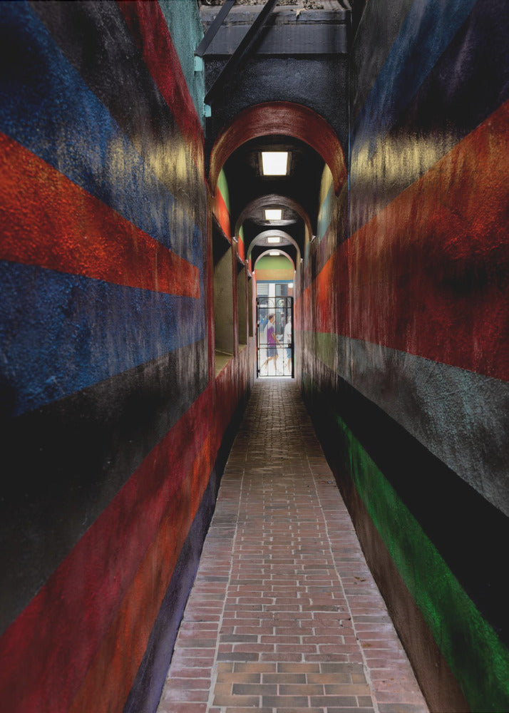 A long, narrow brick alleyway viewed from a one-point perspective, with walls painted in wide, colorful horizontal stripes of red, blue, and black, leading to a distant, sunlit exit where a person stands. Wall Art