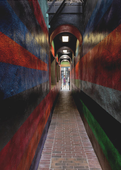 A long, narrow brick alleyway viewed from a one-point perspective, with walls painted in wide, colorful horizontal stripes of red, blue, and black, leading to a distant, sunlit exit where a person stands. Wall Art