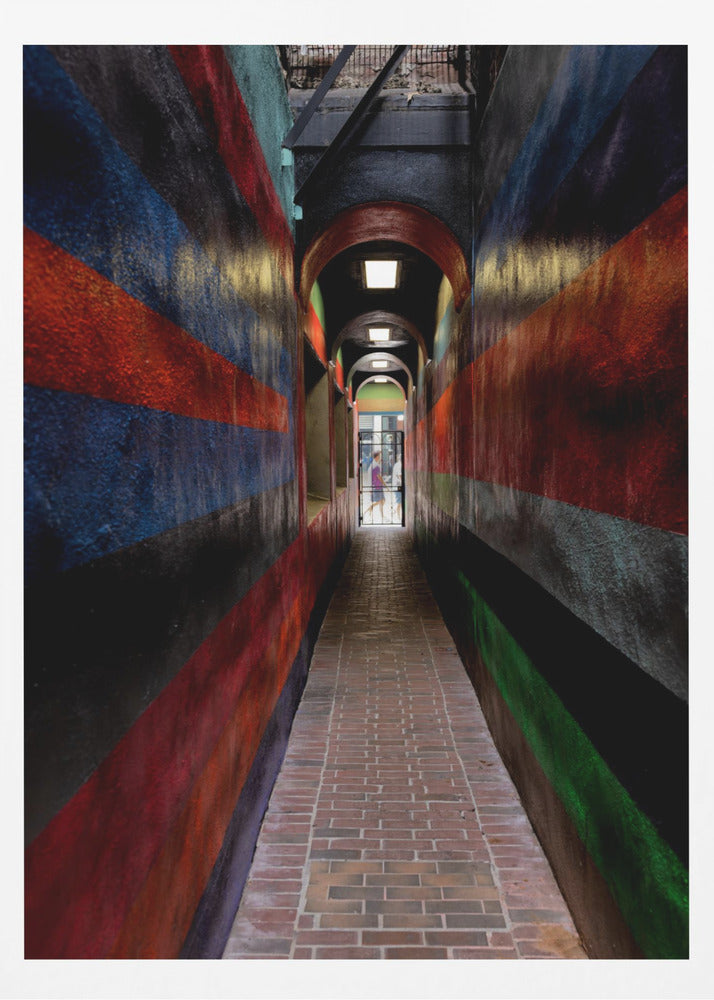 A long, narrow brick alleyway viewed from a one-point perspective, with walls painted in wide, colorful horizontal stripes of red, blue, and black, leading to a distant, sunlit exit where a person stands. Wall Art