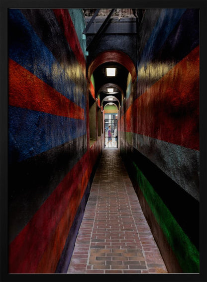 A long, narrow brick alleyway viewed from a one-point perspective, with walls painted in wide, colorful horizontal stripes of red, blue, and black, leading to a distant, sunlit exit where a person stands. Wall Art