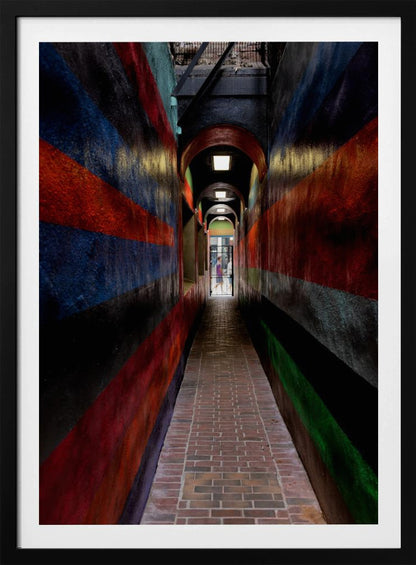 A long, narrow brick alleyway viewed from a one-point perspective, with walls painted in wide, colorful horizontal stripes of red, blue, and black, leading to a distant, sunlit exit where a person stands. Wall Art