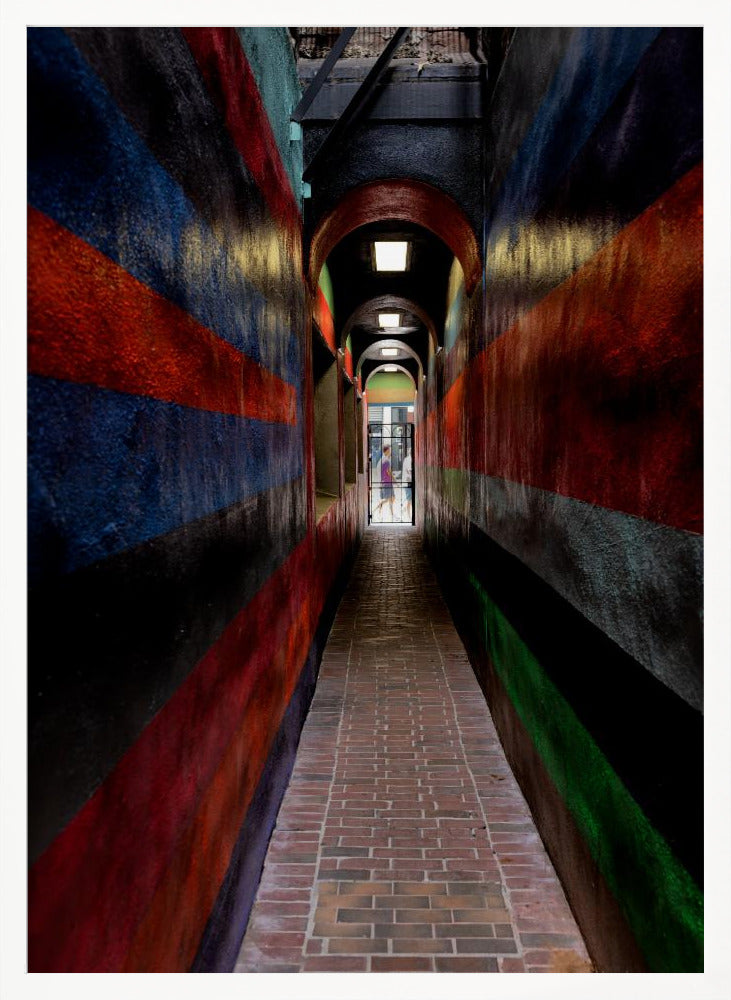 A long, narrow brick alleyway viewed from a one-point perspective, with walls painted in wide, colorful horizontal stripes of red, blue, and black, leading to a distant, sunlit exit where a person stands. Wall Art