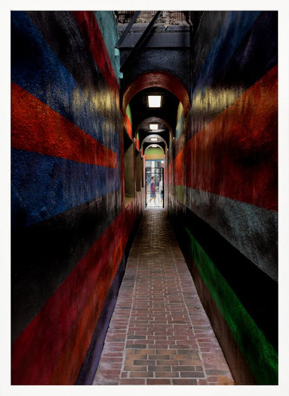 A long, narrow brick alleyway viewed from a one-point perspective, with walls painted in wide, colorful horizontal stripes of red, blue, and black, leading to a distant, sunlit exit where a person stands. Wall Art