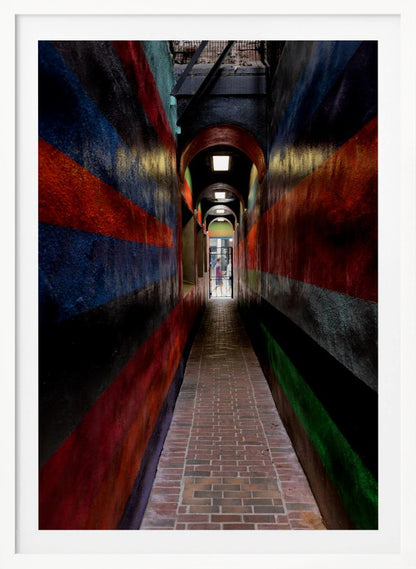 A long, narrow brick alleyway viewed from a one-point perspective, with walls painted in wide, colorful horizontal stripes of red, blue, and black, leading to a distant, sunlit exit where a person stands. Wall Art