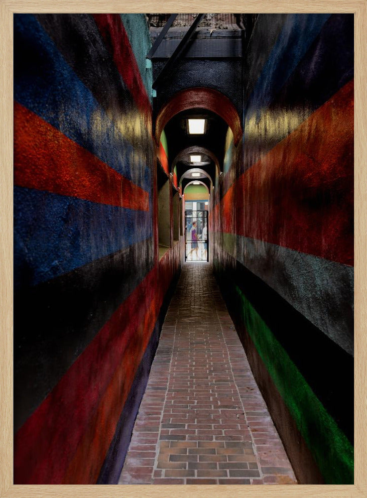 A long, narrow brick alleyway viewed from a one-point perspective, with walls painted in wide, colorful horizontal stripes of red, blue, and black, leading to a distant, sunlit exit where a person stands. Wall Art