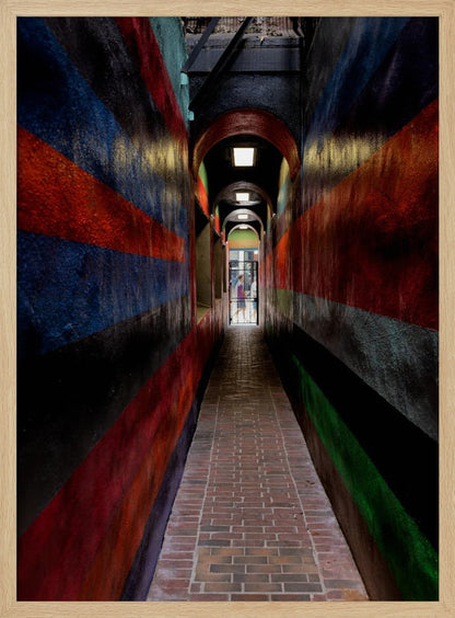 A long, narrow brick alleyway viewed from a one-point perspective, with walls painted in wide, colorful horizontal stripes of red, blue, and black, leading to a distant, sunlit exit where a person stands. Wall Art
