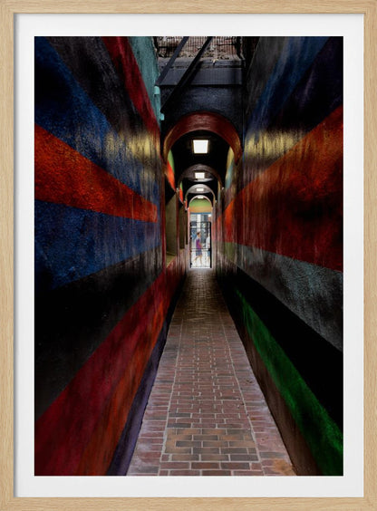 A long, narrow brick alleyway viewed from a one-point perspective, with walls painted in wide, colorful horizontal stripes of red, blue, and black, leading to a distant, sunlit exit where a person stands. Wall Art