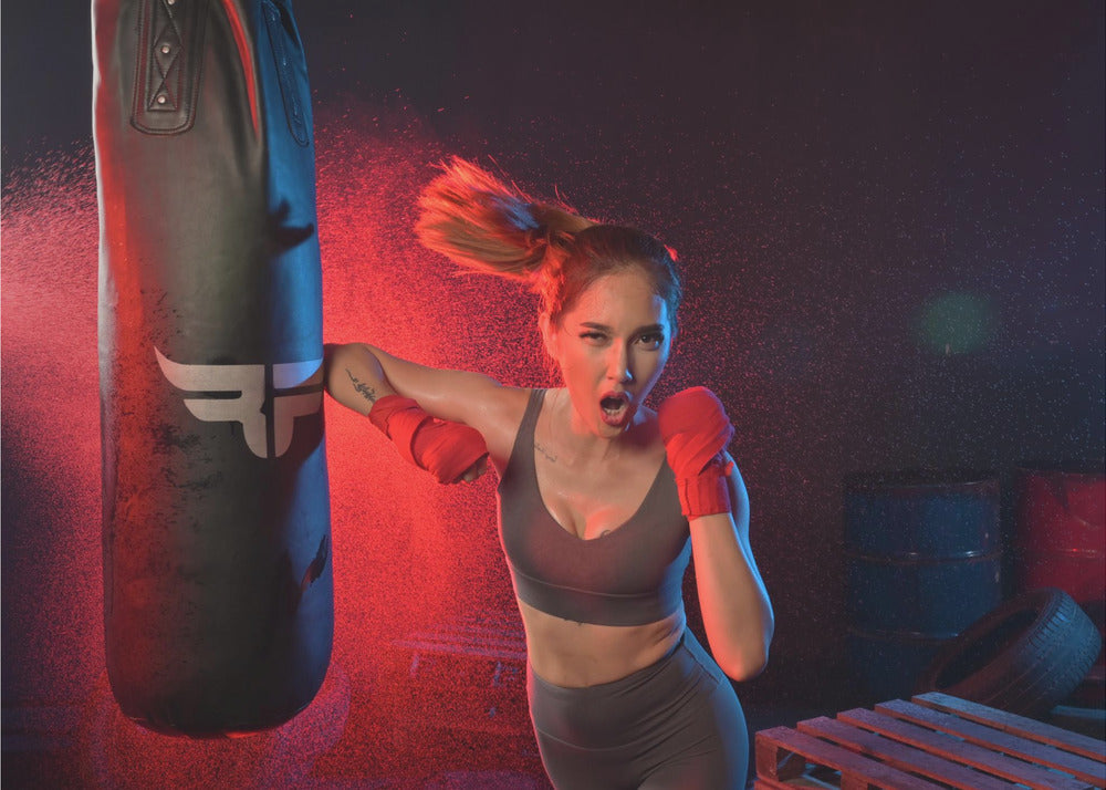 An athletic woman with her hair in a ponytail and red hand wraps fiercely punches a black heavy bag, her mouth open mid-shout. The scene is dramatically lit with red and blue light, and water droplets spray through the air from the impact. Poster