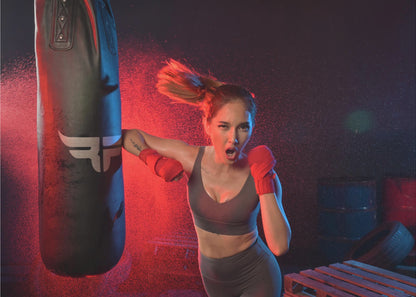 An athletic woman with her hair in a ponytail and red hand wraps fiercely punches a black heavy bag, her mouth open mid-shout. The scene is dramatically lit with red and blue light, and water droplets spray through the air from the impact. Poster
