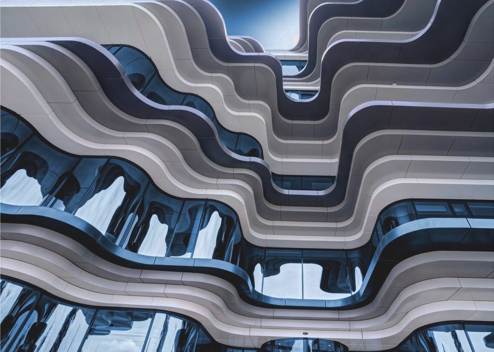 A low-angle view of a modern building with a striking, wavy facade. Alternating bands of light and dark grey create a fluid, ribbon-like pattern, with curved glass windows reflecting the deep blue sky. Artwork