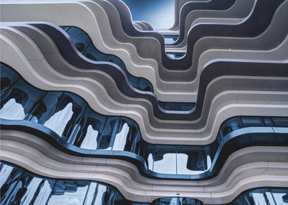 A low-angle view of a modern building with a striking, wavy facade. Alternating bands of light and dark grey create a fluid, ribbon-like pattern, with curved glass windows reflecting the deep blue sky. Artwork
