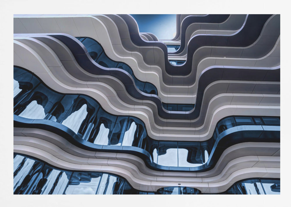 A low-angle view of a modern building with a striking, wavy facade. Alternating bands of light and dark grey create a fluid, ribbon-like pattern, with curved glass windows reflecting the deep blue sky. Artwork