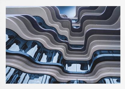 A low-angle view of a modern building with a striking, wavy facade. Alternating bands of light and dark grey create a fluid, ribbon-like pattern, with curved glass windows reflecting the deep blue sky. Artwork