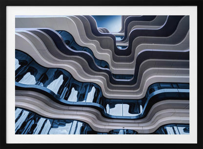A low-angle view of a modern building with a striking, wavy facade. Alternating bands of light and dark grey create a fluid, ribbon-like pattern, with curved glass windows reflecting the deep blue sky. Artwork