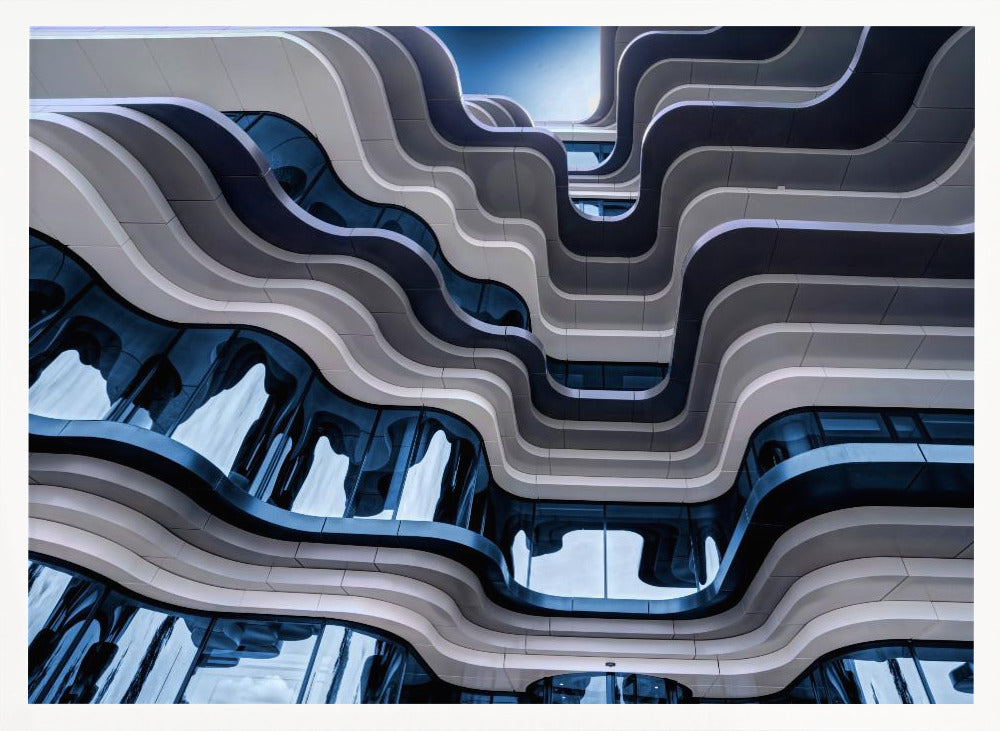 A low-angle view of a modern building with a striking, wavy facade. Alternating bands of light and dark grey create a fluid, ribbon-like pattern, with curved glass windows reflecting the deep blue sky. Artwork
