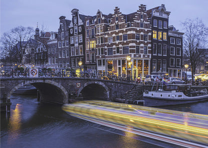 A long-exposure photograph of an Amsterdam canal at dusk, featuring a bright yellow light trail from a passing boat. In the background, illuminated historic canal houses line the water, and an arched stone bridge covered in bicycles crosses the canal. The entire image is presented in a silver frame. Print