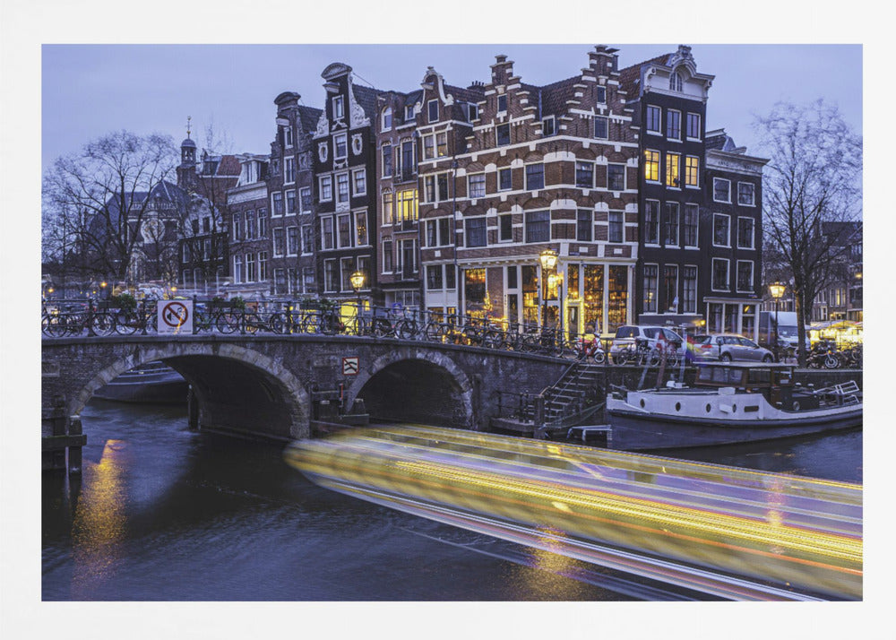 A long-exposure photograph of an Amsterdam canal at dusk, featuring a bright yellow light trail from a passing boat. In the background, illuminated historic canal houses line the water, and an arched stone bridge covered in bicycles crosses the canal. The entire image is presented in a silver frame. Print