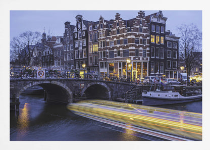 A long-exposure photograph of an Amsterdam canal at dusk, featuring a bright yellow light trail from a passing boat. In the background, illuminated historic canal houses line the water, and an arched stone bridge covered in bicycles crosses the canal. The entire image is presented in a silver frame. Print