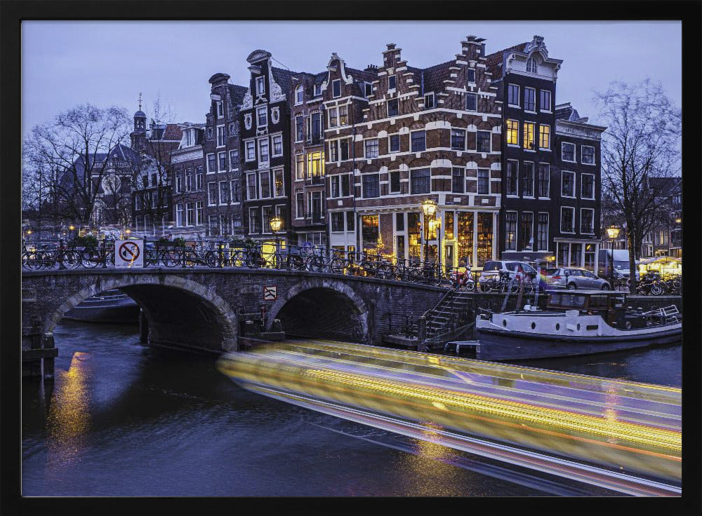 A long-exposure photograph of an Amsterdam canal at dusk, featuring a bright yellow light trail from a passing boat. In the background, illuminated historic canal houses line the water, and an arched stone bridge covered in bicycles crosses the canal. The entire image is presented in a silver frame. Print