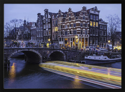 A long-exposure photograph of an Amsterdam canal at dusk, featuring a bright yellow light trail from a passing boat. In the background, illuminated historic canal houses line the water, and an arched stone bridge covered in bicycles crosses the canal. The entire image is presented in a silver frame. Print
