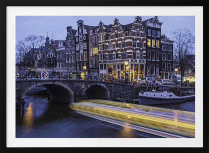 A long-exposure photograph of an Amsterdam canal at dusk, featuring a bright yellow light trail from a passing boat. In the background, illuminated historic canal houses line the water, and an arched stone bridge covered in bicycles crosses the canal. The entire image is presented in a silver frame. Print