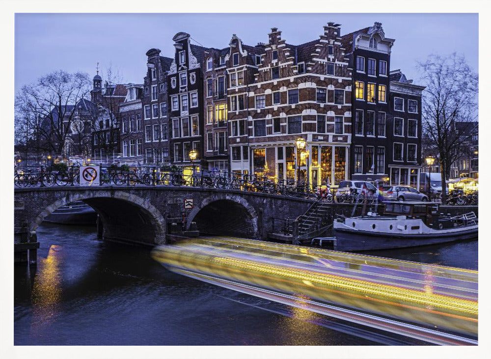 A long-exposure photograph of an Amsterdam canal at dusk, featuring a bright yellow light trail from a passing boat. In the background, illuminated historic canal houses line the water, and an arched stone bridge covered in bicycles crosses the canal. The entire image is presented in a silver frame. Print