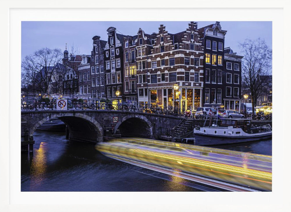 A long-exposure photograph of an Amsterdam canal at dusk, featuring a bright yellow light trail from a passing boat. In the background, illuminated historic canal houses line the water, and an arched stone bridge covered in bicycles crosses the canal. The entire image is presented in a silver frame. Print