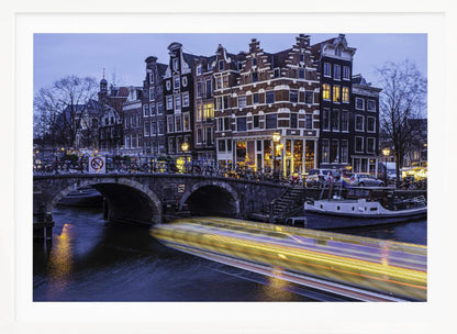 A long-exposure photograph of an Amsterdam canal at dusk, featuring a bright yellow light trail from a passing boat. In the background, illuminated historic canal houses line the water, and an arched stone bridge covered in bicycles crosses the canal. The entire image is presented in a silver frame. Print
