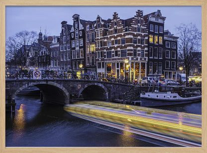 A long-exposure photograph of an Amsterdam canal at dusk, featuring a bright yellow light trail from a passing boat. In the background, illuminated historic canal houses line the water, and an arched stone bridge covered in bicycles crosses the canal. The entire image is presented in a silver frame. Print
