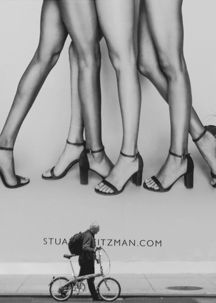 A black and white street photograph showing a stark contrast between a large fashion advertisement and a person on the sidewalk. The ad features multiple pairs of long female legs wearing high-heeled sandals. Below the ad, an older man with a backpack walks by, pushing a small foldable bicycle. Artwork