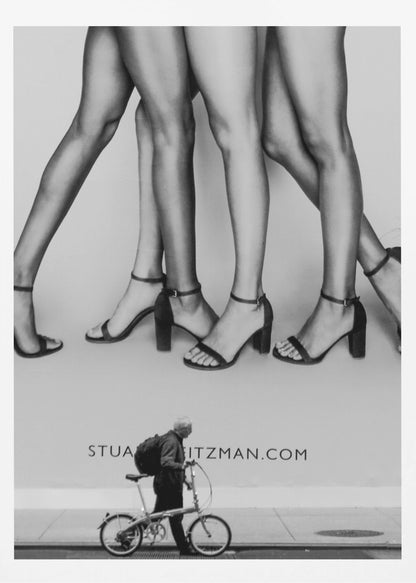 A black and white street photograph showing a stark contrast between a large fashion advertisement and a person on the sidewalk. The ad features multiple pairs of long female legs wearing high-heeled sandals. Below the ad, an older man with a backpack walks by, pushing a small foldable bicycle. Artwork