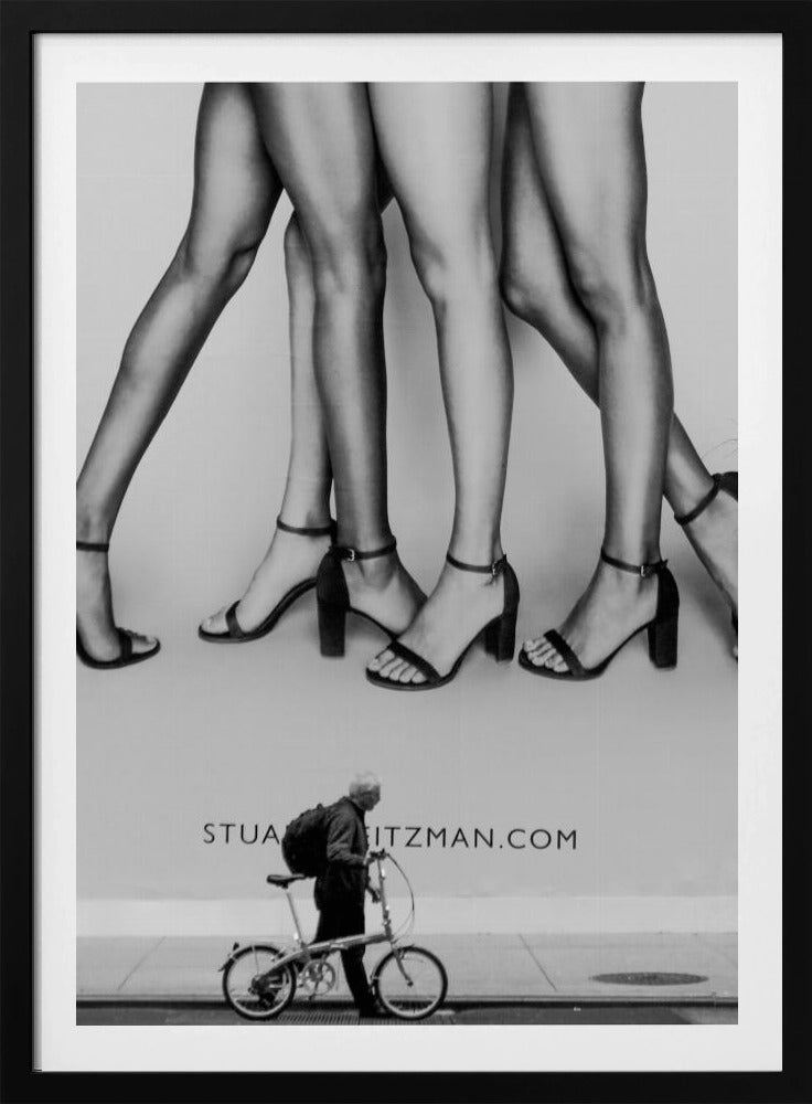 A black and white street photograph showing a stark contrast between a large fashion advertisement and a person on the sidewalk. The ad features multiple pairs of long female legs wearing high-heeled sandals. Below the ad, an older man with a backpack walks by, pushing a small foldable bicycle. Artwork