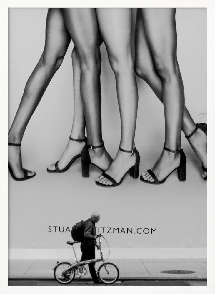 A black and white street photograph showing a stark contrast between a large fashion advertisement and a person on the sidewalk. The ad features multiple pairs of long female legs wearing high-heeled sandals. Below the ad, an older man with a backpack walks by, pushing a small foldable bicycle. Artwork