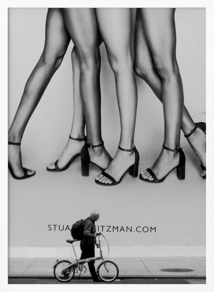 A black and white street photograph showing a stark contrast between a large fashion advertisement and a person on the sidewalk. The ad features multiple pairs of long female legs wearing high-heeled sandals. Below the ad, an older man with a backpack walks by, pushing a small foldable bicycle. Artwork