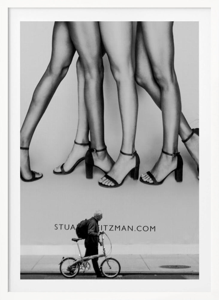 A black and white street photograph showing a stark contrast between a large fashion advertisement and a person on the sidewalk. The ad features multiple pairs of long female legs wearing high-heeled sandals. Below the ad, an older man with a backpack walks by, pushing a small foldable bicycle. Artwork