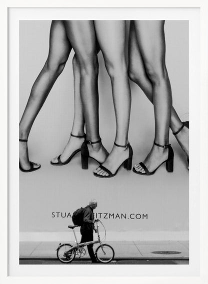 A black and white street photograph showing a stark contrast between a large fashion advertisement and a person on the sidewalk. The ad features multiple pairs of long female legs wearing high-heeled sandals. Below the ad, an older man with a backpack walks by, pushing a small foldable bicycle. Artwork