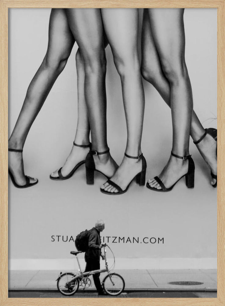 A black and white street photograph showing a stark contrast between a large fashion advertisement and a person on the sidewalk. The ad features multiple pairs of long female legs wearing high-heeled sandals. Below the ad, an older man with a backpack walks by, pushing a small foldable bicycle. Artwork