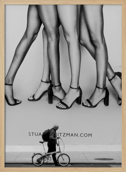 A black and white street photograph showing a stark contrast between a large fashion advertisement and a person on the sidewalk. The ad features multiple pairs of long female legs wearing high-heeled sandals. Below the ad, an older man with a backpack walks by, pushing a small foldable bicycle. Artwork