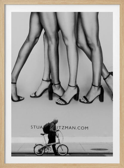A black and white street photograph showing a stark contrast between a large fashion advertisement and a person on the sidewalk. The ad features multiple pairs of long female legs wearing high-heeled sandals. Below the ad, an older man with a backpack walks by, pushing a small foldable bicycle. Artwork