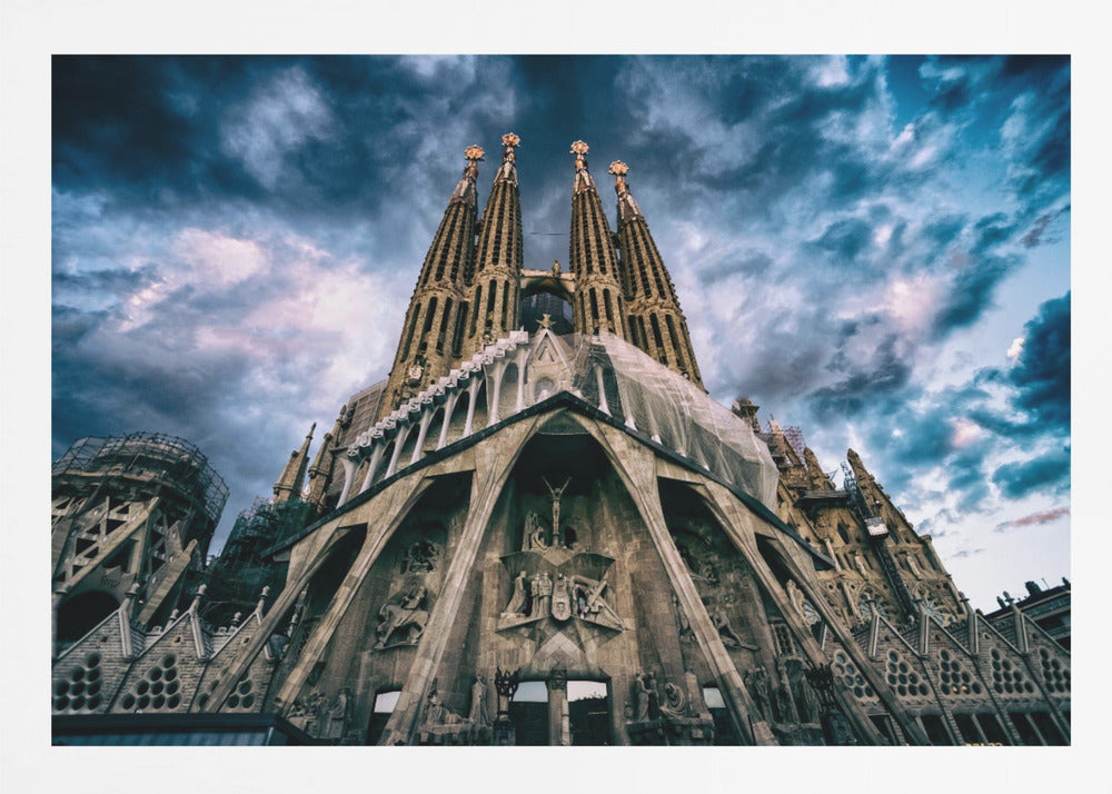 A dramatic, low-angle photograph of the Sagrada Familia in Barcelona, with its ornate stone facade and towering spires reaching up into a turbulent, dark blue and cloudy sky. The perspective emphasizes the grand scale and intricate detail of Gaudí's architecture. Print