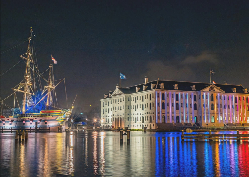 A vibrant nighttime photograph of the National Maritime Museum and a replica of the East Indiaman Amsterdam ship in a harbor. Both are illuminated with colorful lights that cast beautiful reflections on the dark water. Wall Art