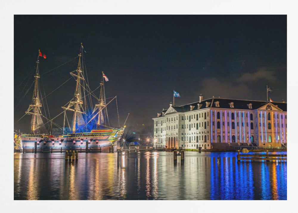 A vibrant nighttime photograph of the National Maritime Museum and a replica of the East Indiaman Amsterdam ship in a harbor. Both are illuminated with colorful lights that cast beautiful reflections on the dark water. Wall Art