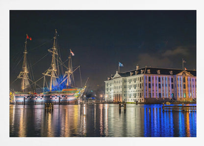 A vibrant nighttime photograph of the National Maritime Museum and a replica of the East Indiaman Amsterdam ship in a harbor. Both are illuminated with colorful lights that cast beautiful reflections on the dark water. Wall Art