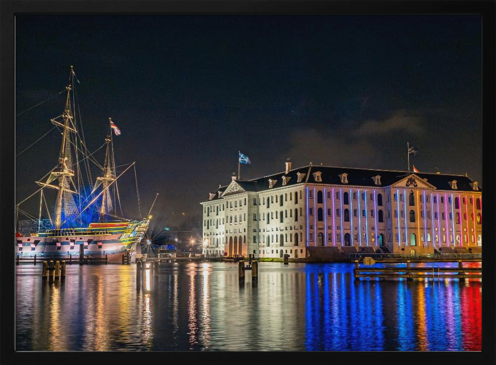 A vibrant nighttime photograph of the National Maritime Museum and a replica of the East Indiaman Amsterdam ship in a harbor. Both are illuminated with colorful lights that cast beautiful reflections on the dark water. Wall Art