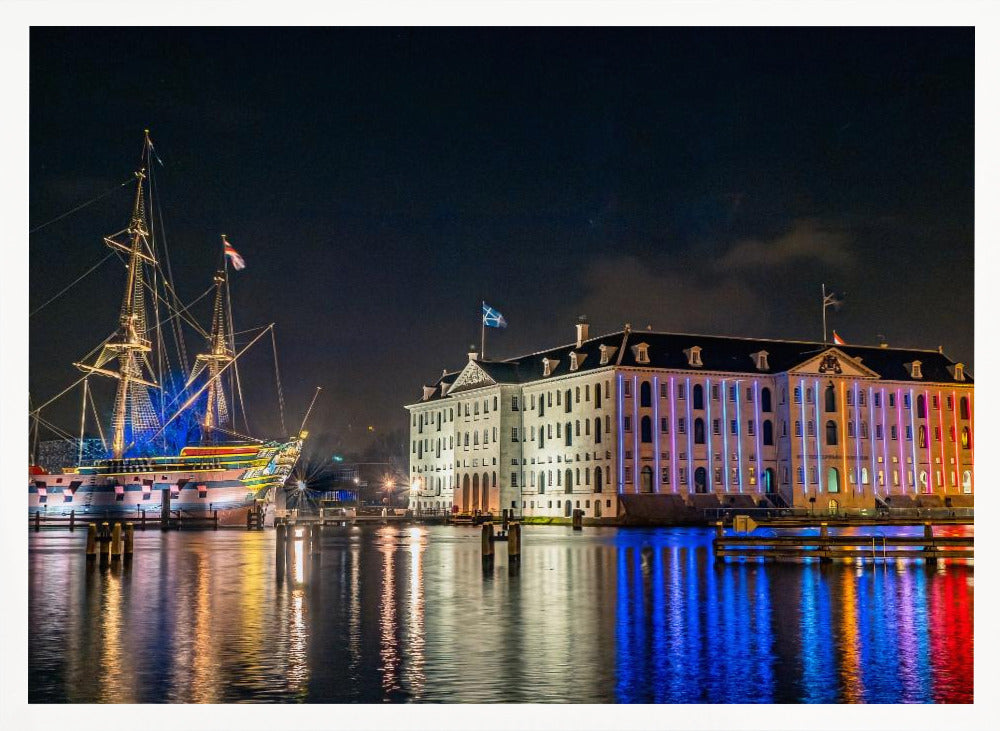 A vibrant nighttime photograph of the National Maritime Museum and a replica of the East Indiaman Amsterdam ship in a harbor. Both are illuminated with colorful lights that cast beautiful reflections on the dark water. Wall Art