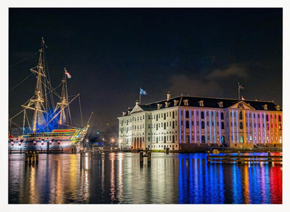 A vibrant nighttime photograph of the National Maritime Museum and a replica of the East Indiaman Amsterdam ship in a harbor. Both are illuminated with colorful lights that cast beautiful reflections on the dark water. Wall Art