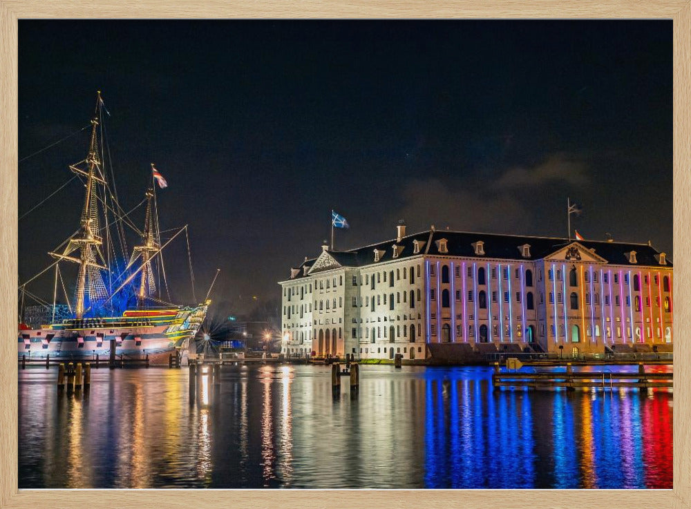 A vibrant nighttime photograph of the National Maritime Museum and a replica of the East Indiaman Amsterdam ship in a harbor. Both are illuminated with colorful lights that cast beautiful reflections on the dark water. Wall Art