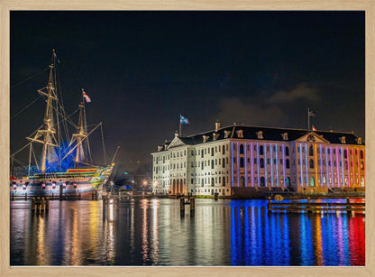 A vibrant nighttime photograph of the National Maritime Museum and a replica of the East Indiaman Amsterdam ship in a harbor. Both are illuminated with colorful lights that cast beautiful reflections on the dark water. Wall Art