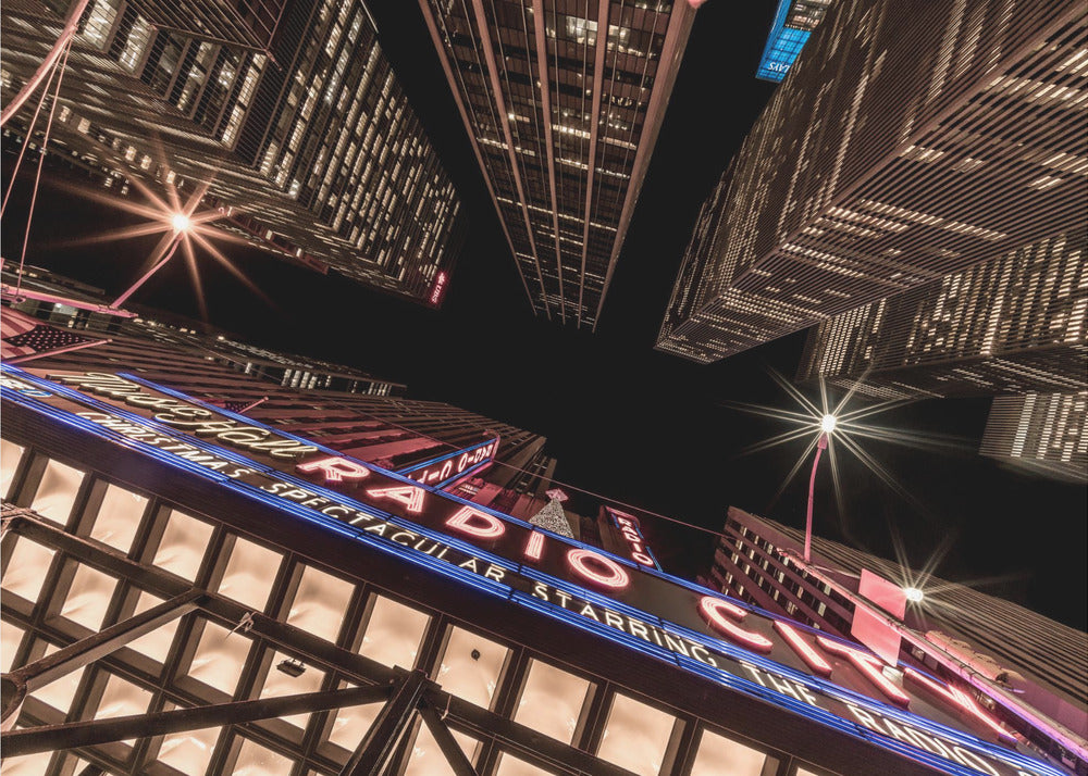 A dramatic low-angle shot looking up at the iconic Radio City Music Hall marquee at night, surrounded by towering, illuminated skyscrapers against a dark sky. Decor