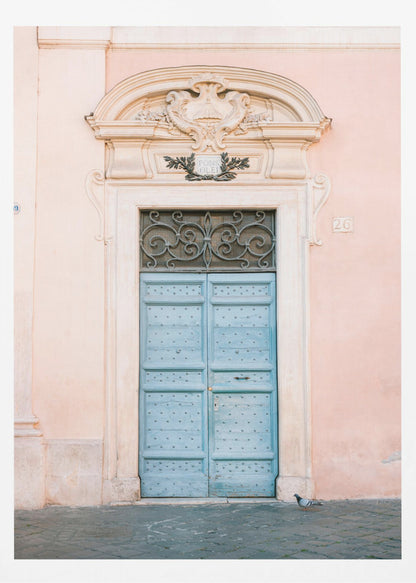 A light blue, studded double door set into a pale pink wall. The doorway is framed in white stone with an ornate pediment and a decorative wrought-iron transom window above the door. A single pigeon stands on the cobblestone pavement in front. Print